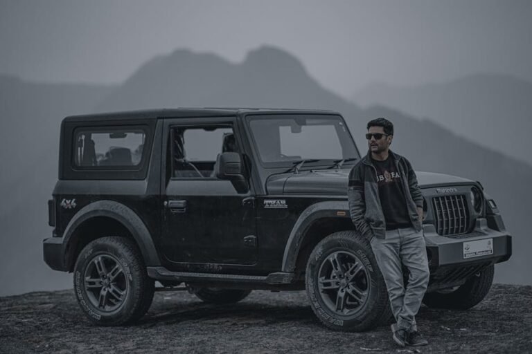 A man in a black jacket and sunglasses leans on a Mahindra 4x4 jeep, set against a misty mountain landscape.