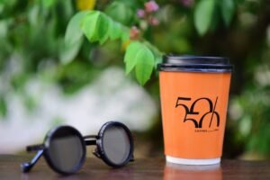 Orange coffee cup and trendy sunglasses on a table outdoors, lush green backdrop.