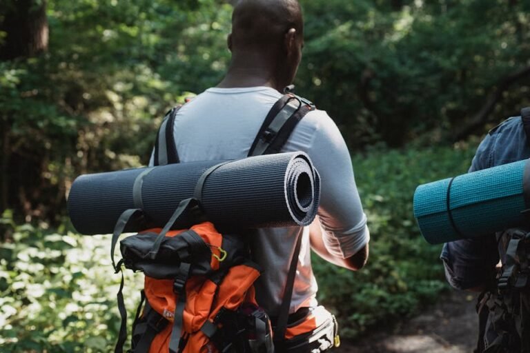 Two men hiking through a verdant forest, carrying backpacks and yoga mats on a sunny day.
