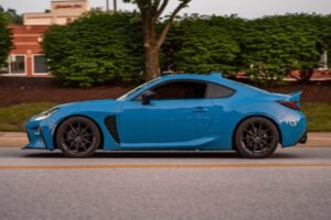 A modern blue sports car speeding on a suburban street with greenery and buildings in the background.