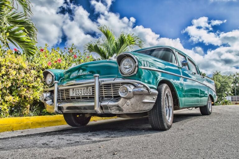 A beautifully restored classic car on a sunny road in Havana, Cuba, with lush foliage and a vibrant sky.