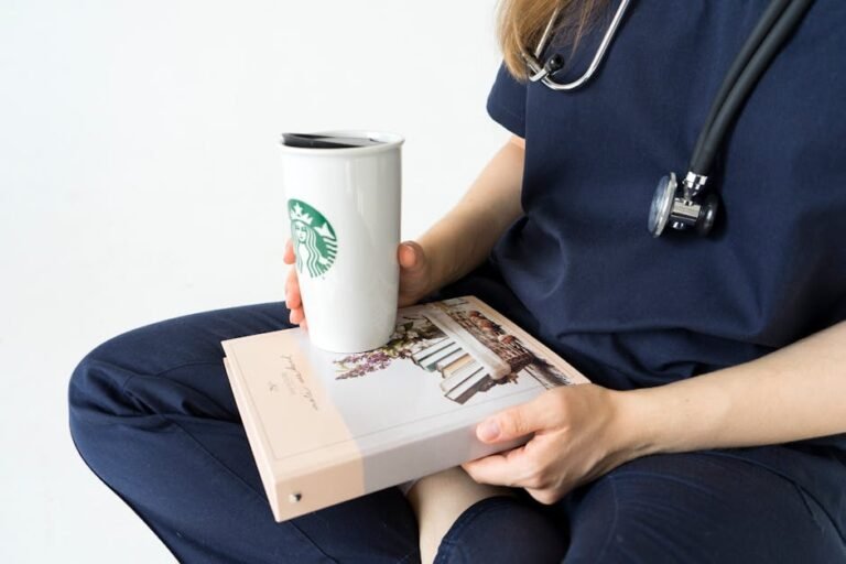 A nurse in scrubs holds a coffee cup and book while sitting indoors.