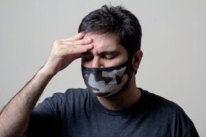 A man in a black shirt wears a camo face mask, showing signs of headache and illness.