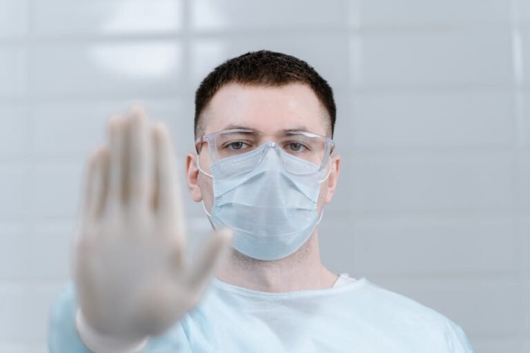 Healthcare worker wearing protective gear and mask making stop gesture in hospital setting.
